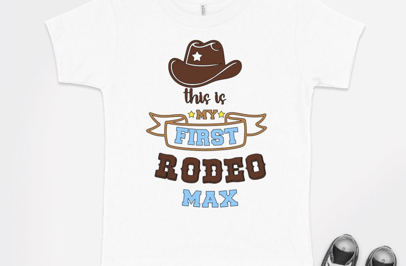 Pode incluir: Camiseta branca com um chap&eacute;u de cowboy marrom, uma faixa marrom com o texto "This is my first rodeo Max" em letras amarelas, azuis e marrons.