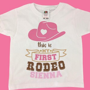 Pode incluir: Camiseta branca com um chap&eacute;u de cowboy rosa, uma faixa dourada com o texto "This is my first rodeo" e o nome "Sienna" em rosa.