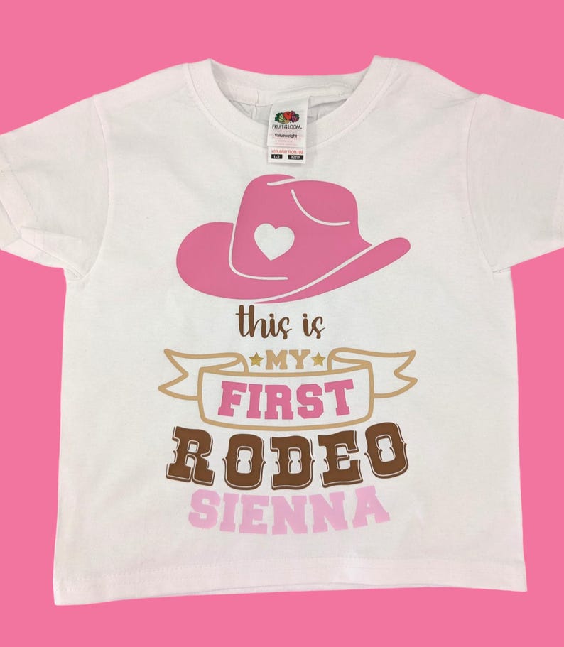 Pode incluir: Camiseta branca com um chap&eacute;u de cowboy rosa, uma faixa dourada com o texto "This is my first rodeo" e o nome "Sienna" em rosa.