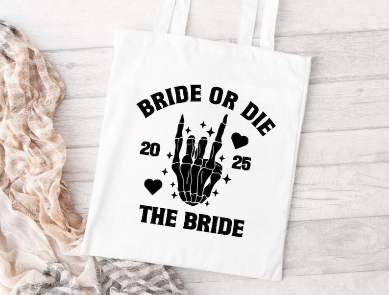 May include: White tote bag with black text and graphics. The text reads "BRIDE OR DIE" above a black skeleton hand making a rock and roll gesture. Below the hand is the text "THE BRIDE", with the numbers "20" and "25" on either side.