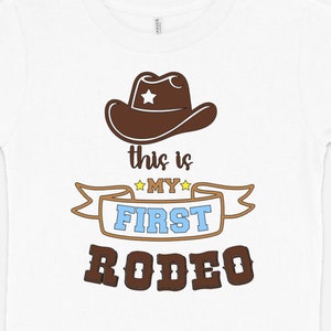 Pode incluir: Camiseta branca com um chap&eacute;u de cowboy marrom, uma faixa marrom com o texto "This is my first rodeo" em letras amarelas, azuis e marrons.
