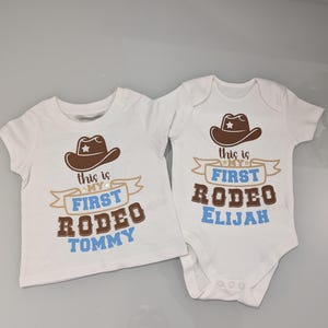 Pode incluir: Dois bodies brancos para beb&ecirc; com chap&eacute;us de cowboy marrons e texto azul. Um body diz "This is my first rodeo Tommy" e o outro diz "This is my first rodeo Elijah".