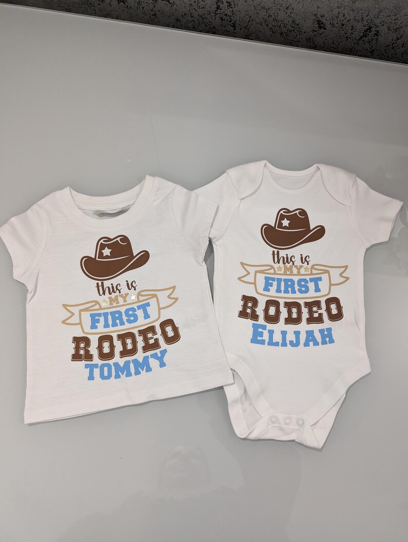 Pode incluir: Dois bodies brancos para beb&ecirc; com chap&eacute;us de cowboy marrons e texto azul. Um body diz "This is my first rodeo Tommy" e o outro diz "This is my first rodeo Elijah".