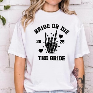 May include: White t-shirt with a black graphic that reads "BRIDE OR DIE" above a skeleton hand making a rock-and-roll gesture. The text "20 25" and "THE BRIDE" are also in black. Two black hearts and stars complete the design.