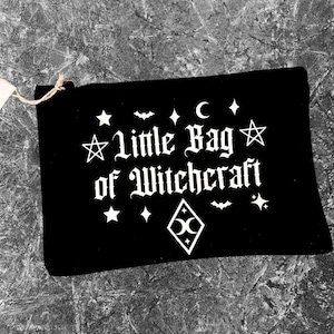 Puede incluir: Bolsa rectangular negra con el texto "Little Bag of Witchcraft" en blanco. Decorada con estrellas, diamantes, una luna creciente y un murciélago blancos. Una pequeña etiqueta está adjunta.