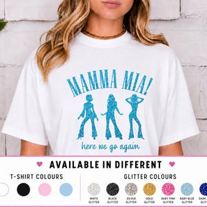 May include: A white short-sleeved t-shirt featuring a blue glitter graphic of three figures in flared trousers. The text reads MAMMA MIA! in large blue glitter letters above the figures and here we go again in smaller blue text below them.