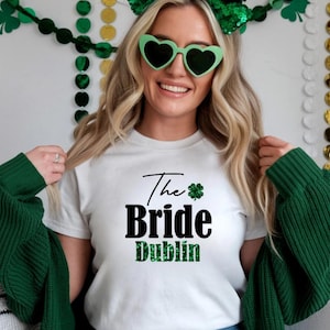 May include: White t-shirt with the text "The Bride Dublin" in black and green. The shirt features a shamrock graphic. The person in the image is wearing green heart-shaped sunglasses and a green headband, with a green sweater draped over their shoulders.