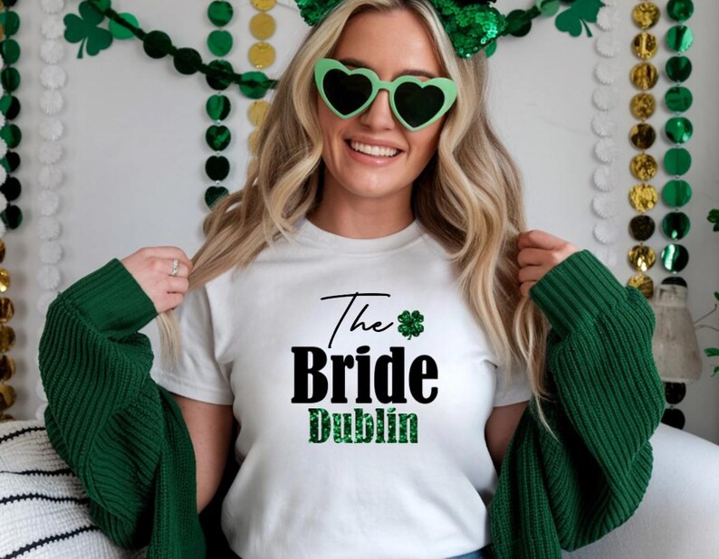 May include: White t-shirt with the text "The Bride Dublin" in black and green. The shirt features a shamrock graphic. The person in the image is wearing green heart-shaped sunglasses and a green headband, with a green sweater draped over their shoulders.