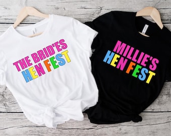 Pode incluir: Duas camisetas, uma branca e outra preta, com texto colorido. A camiseta branca diz "THE BRID'ES HEN FEST" e a preta "MILLIE'S HEN FEST". Ambas estão penduradas em cabides de madeira.