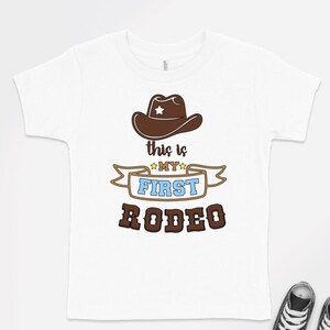 Pode incluir: Camiseta branca com um chap&eacute;u de cowboy marrom e o texto "This is my first rodeo" em marrom, azul e amarelo.