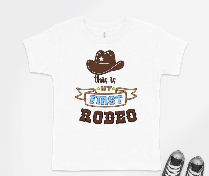 Pode incluir: Camiseta branca com um chap&eacute;u de cowboy marrom e o texto "This is my first rodeo" em marrom, azul e amarelo.