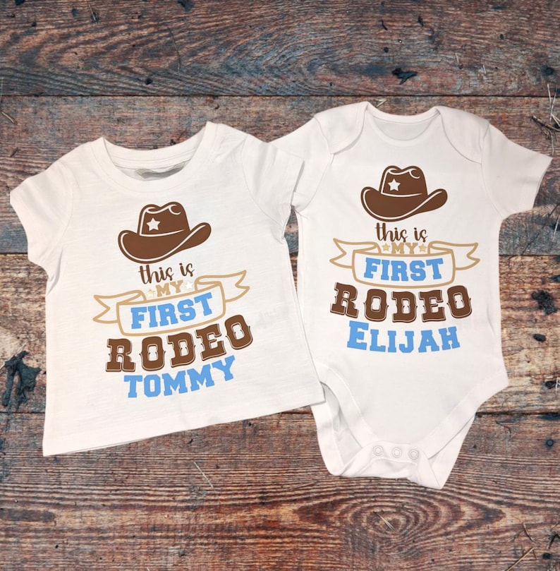 Pode incluir: Duas camisetas brancas com chap&eacute;us de cowboy marrons e texto azul que diz "This is my first rodeo" com um nome embaixo. Uma camiseta diz "Tommy" e a outra diz "Elijah".