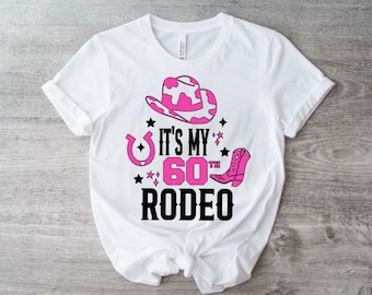 Pode incluir: Camiseta branca com um chapéu de cowboy rosa, uma bota de cowboy rosa e o texto "It's My 60th Rodeo" em preto.