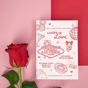 May include: A red rose with green leaves next to a hen party invitation. The invitation is white with red illustrations and text, including "Lucky in Love" and "Welcome to Las Vegas". The background is pink with a red card.