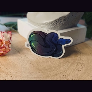 Peut inclure: Un autocollant représentant un serpent bleu iridescent enroulé avec un dégradé vert et violet. Le serpent est enroulé en spirale, la tête tournée vers la droite de l'autocollant.
