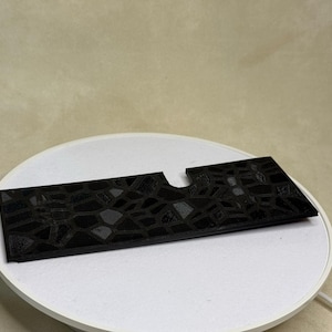 Puede incluir: Un objeto rectangular negro con un patr&oacute;n geom&eacute;trico texturizado y un recorte curvo. El objeto se muestra sobre una superficie circular blanca, lo que sugiere una presentaci&oacute;n o exhibici&oacute;n del producto.