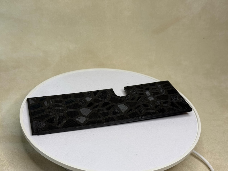 Puede incluir: Un objeto rectangular negro con un patr&oacute;n geom&eacute;trico texturizado y un recorte curvo. El objeto se muestra sobre una superficie circular blanca, lo que sugiere una presentaci&oacute;n o exhibici&oacute;n del producto.