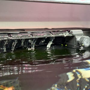 Könnte beinhalten: Ein schwarzer Plastik-Wasserfallfilter für ein Aquarium. Der Filter hat eine Reihe kleiner, rechteckiger Öffnungen, die den Wasserfluss ermöglichen. Das Wasser fließt über den Filter und in das Aquarium darunter.