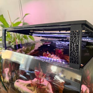 May include: A black aquarium with a decorative patterned frame. The tank is filled with water and contains plants and decorative elements. The top of the tank has a black frame with a built-in light fixture.