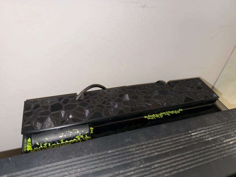 Puede incluir: Un filtro de acuario negro con una tapa texturizada y un asa. El filtro tiene una secci&oacute;n con vegetaci&oacute;n verde visible. El filtro est&aacute; dise&ntilde;ado para mantener el agua limpia para la vida acu&aacute;tica.