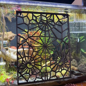 May include: A dark brown, geometric patterned screen, likely for an aquarium, is the focal point. The screen has a complex, symmetrical design with circular and angular shapes. The background reveals an aquarium with plants and a decorative skull.