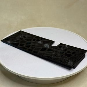 Puede incluir: Un objeto rectangular negro con un patr&oacute;n texturizado similar a un mosaico. Tiene un recorte en un lado y est&aacute; colocado sobre una plataforma blanca y circular. El fondo es de color beige neutro.