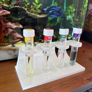 May include: A white plastic test tube holder with four clear glass tubes. Each tube has a different coloured cap: yellow, red, green, and purple. The tubes contain liquids of varying colours, and the holder has the letters "MMS" on it.