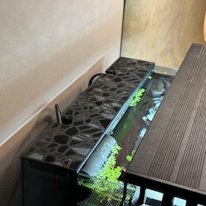 Puede incluir: Un acuario negro con una tapa estampada y una cubierta ranurada separada. El tanque contiene agua y plantas verdes. Un peque&ntilde;o tubo plateado se extiende desde la parte superior estampada. El acuario est&aacute; colocado contra una pared de color neutro.