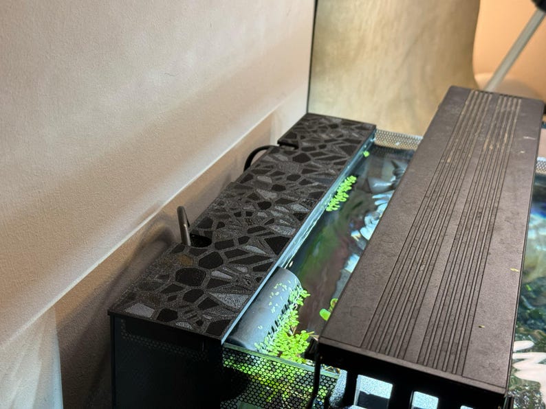 Puede incluir: Un acuario negro con una tapa estampada y una cubierta ranurada separada. El tanque contiene agua y plantas verdes. Un peque&ntilde;o tubo plateado se extiende desde la parte superior estampada. El acuario est&aacute; colocado contra una pared de color neutro.