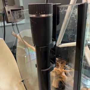 May include: A black aquarium heater with a temperature dial and mounting brackets. The heater is cylindrical and partially submerged in a glass aquarium. The aquarium contains driftwood and other aquatic elements.