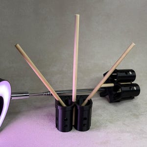 May include: A black plastic holder with three slots for holding wooden dowels. The dowels are arranged in a fan shape. The holder is sitting on a gray surface.