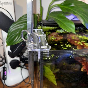 Könnte beinhalten: Ein klarer Acryl-Aquarium-Werkzeughalter mit Edelstahlscheren und einem Metallschaber. Der Halter ist an der Seite eines Glasaquariums befestigt, in dem grüne Pflanzen und Wasser sichtbar sind. Im Hintergrund befindet sich ein weißer Überspannungsschutz.