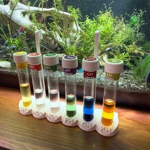 May include: A white test tube rack holding six glass tubes with coloured liquids. Each tube has a white cap with a label: NH2, NO2, NO3, pH, GH, and KH. The tubes are in front of an aquarium.