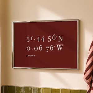 May include: A rectangular wall art print in a silver frame featuring a deep red background with white text displaying the coordinates 51.44 56 N and 0.06 76 W, followed by the word LONDON in a clean, sans-serif font centred on the print.