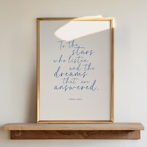 May include: A framed print with the quote "To the stars who listen, and the dreams that are answered." in blue script. The print is in a gold frame and rests on a wooden shelf. The author's name, Sarah J. Maas, is at the bottom.