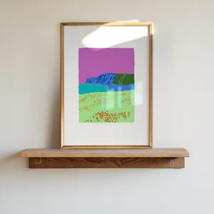 May include: A framed landscape art print with a gold frame. The artwork features a vibrant color palette, including a purple sky, blue mountains, and green and yellow fields. The print is displayed on a wooden shelf.