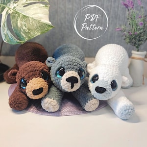 May include: Three plush crochet bears in brown, gray, and white, each featuring large glossy eyes and a stitched nose. The bears are positioned side-by-side, with a circular graphic overlay in the upper center containing the text PDF Pattern.