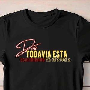 May include: Black t-shirt with the Spanish text "Dios Todavia Esta Escribiendo Tu Historia" in a stylized design. The text is in red, yellow, and white, with a cursive font for "Dios".