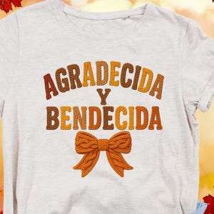 May include: A light gray t-shirt with the words "AGRADECIDA Y BENDECIDA" in brown and orange embroidered letters. A bow design is below the text. The background has autumn leaves.