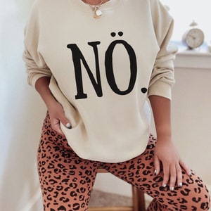 May include: A beige sweatshirt with the word "N&Ouml;." printed in black on the front. The model is wearing leopard print leggings.