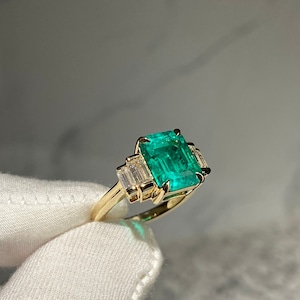 May include: A gold ring featuring a large emerald green gemstone and smaller baguette-cut diamonds on the band.