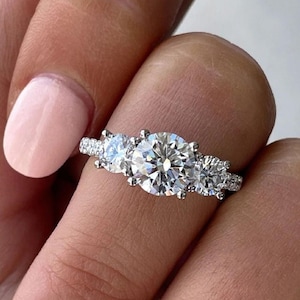 May include: A silver three-stone engagement ring with a large round centre diamond and two smaller round diamonds on either side.