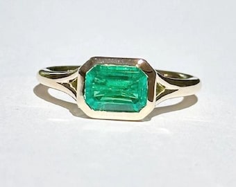 May include: A gold ring with an emerald green gemstone set in a square bezel. The ring has a simple, elegant design.
