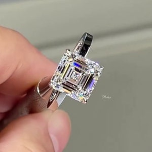May include: A silver ring with a large, square-shaped, clear diamond. The diamond is set in a halo of smaller diamonds. The ring is on a finger.