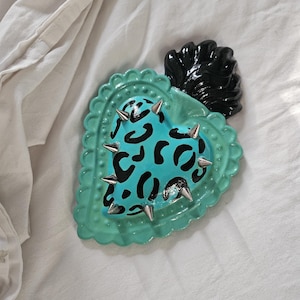 May include: A heart-shaped wall ornament featuring a bright turquoise center with a black leopard print pattern and silver metal spikes. The heart is framed by a scalloped turquoise border and topped with a glossy black flame-like accent.