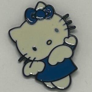 May include: A white enamel pin shaped like a cartoon cat character with black outlines. The character wears a dark blue dress and a matching dark blue bow on its head. It has black eyes, a yellow nose, and black whiskers on both sides of its face.