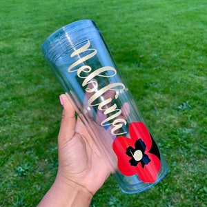 Op de afbeelding: Een doorzichtige plastic tumbler met een gouden vinyl ontwerp met de tekst "Melina" en een rode klaproos afbeelding.