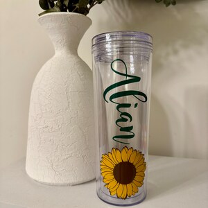Può includere: Un bicchiere di plastica trasparente con un design di girasole verde e nero e il nome "Alian" scritto in corsivo verde.
