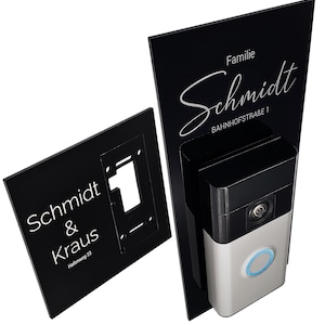 Könnte beinhalten: Schwarzes Türklingelsystem mit einer silbernen und schwarzen Kameraeinheit. Das System umfasst eine Montageplatte und ein Schild mit dem Namen "Schmidt" und Adressinformationen. Der Klingelknopf hat einen blau leuchtenden Ring.