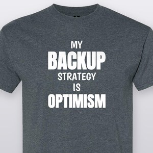 May include: A dark heather gray short-sleeved t-shirt featuring the white text MY BACKUP STRATEGY IS OPTIMISM printed in a bold, sans-serif font across the chest. The garment has a classic crew neckline and a standard fit with short sleeves.